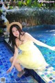 A woman in a yellow dress and straw hat sitting on the edge of a pool.