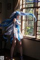 A woman with long blue hair standing in front of a window.